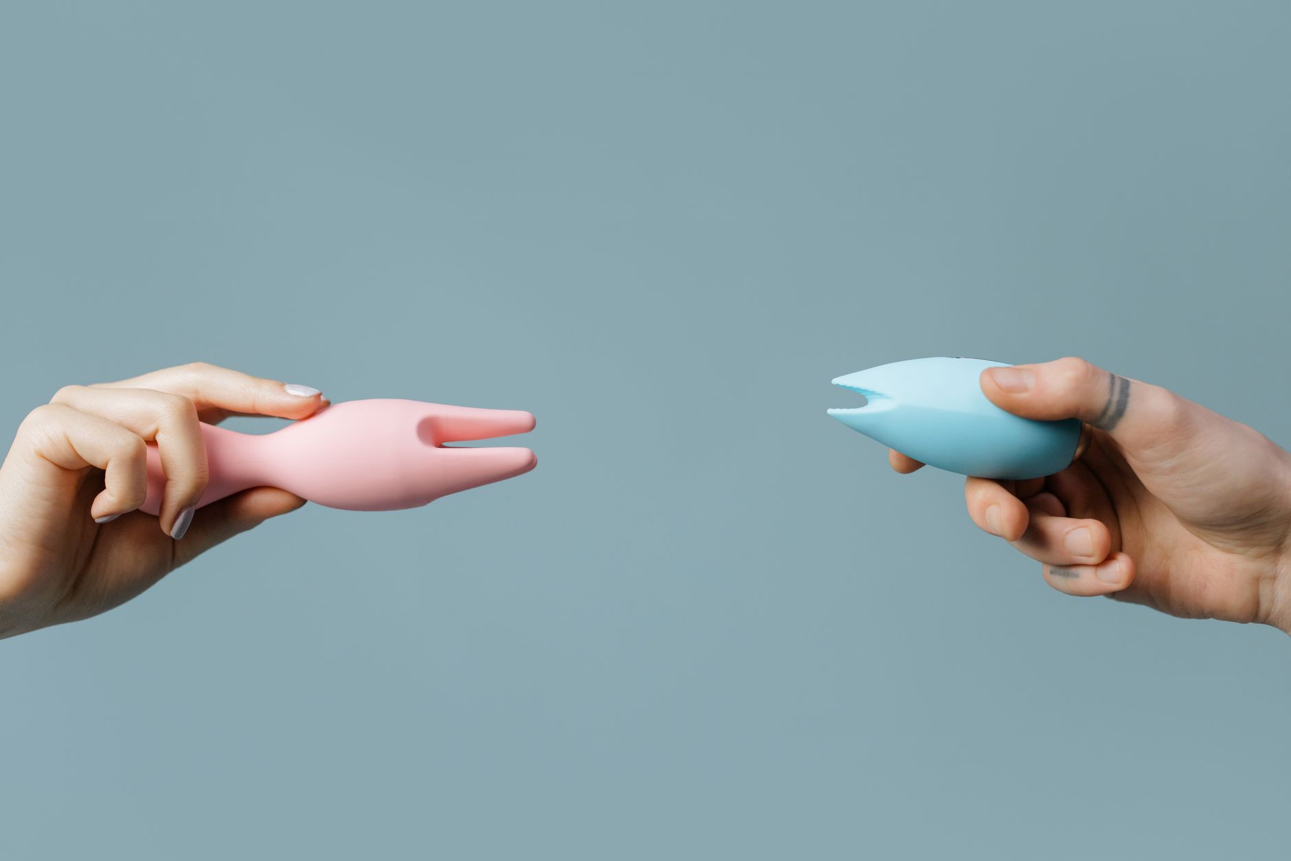 Two hands holding vibrators against a pastel background, representing shared intimacy and couple exploration.